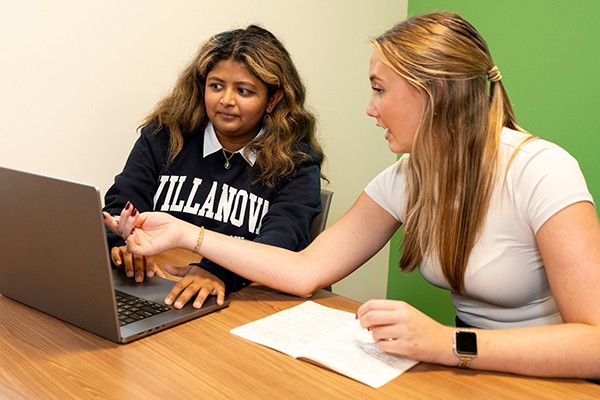 Mother Cabrini Institute on Immigration students sitting together at a table, one has a laptop, the other is writing with a pen and notepad