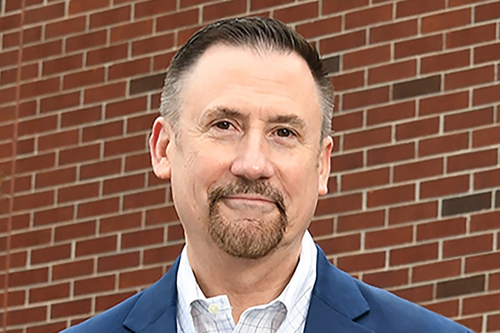 Headshot of Michael Gaynor standing in front of a brick wall wearing a blue suit jacket with a  Villanova University spirit lapel pin.