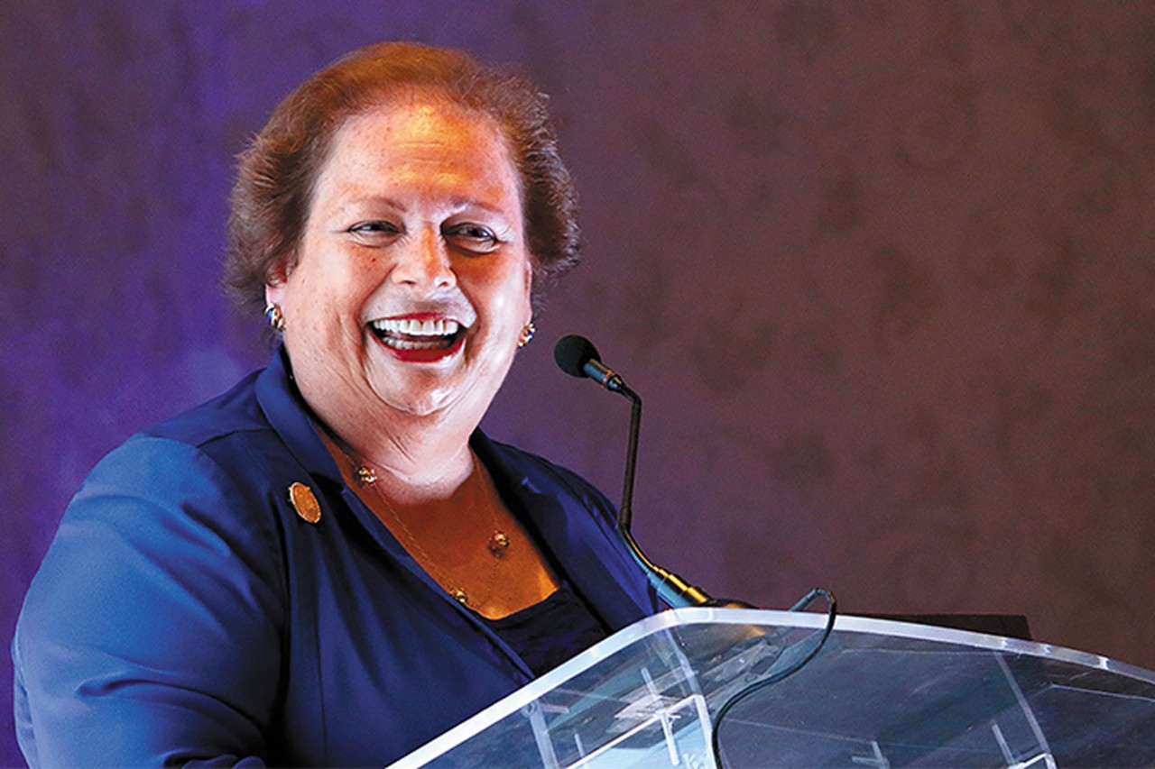 Mari Carmen Aponte dressed in blue standing in front of a podium with a microphone