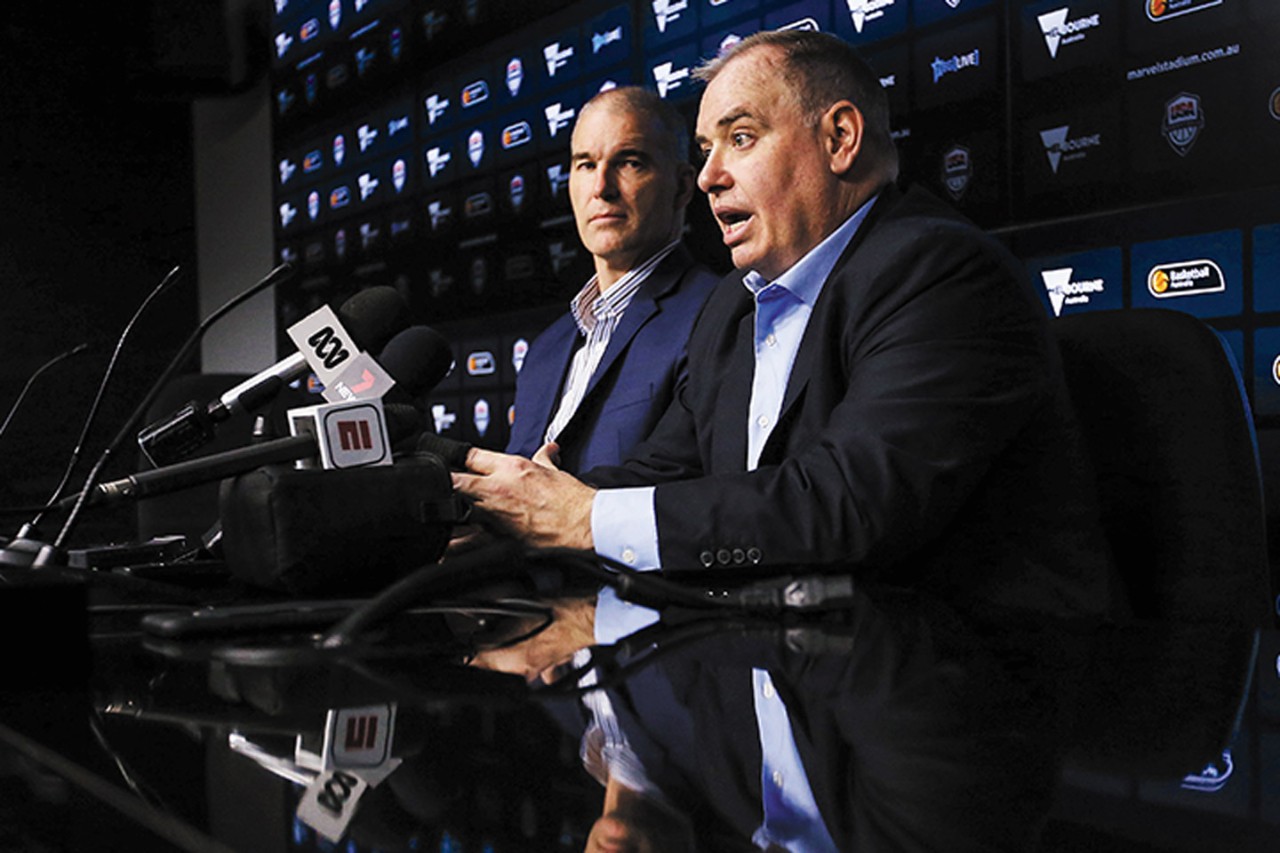 a seated Sean Ford wearing a dark blazer speaks to the media 